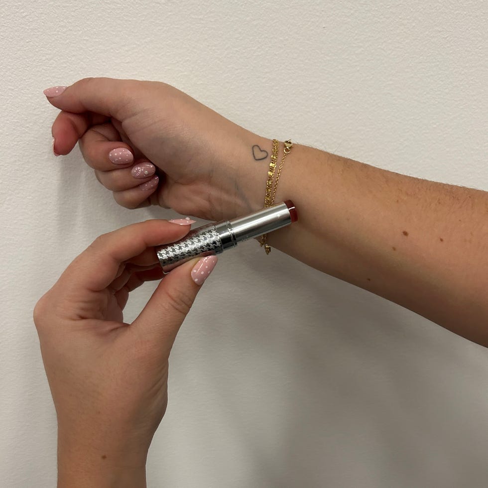 mão segurando um tubo de batom perto de um pulso com uma tatuagem e pulseira