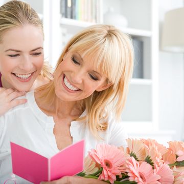 smiling young adult daughter looking over smiling mother's shoulder, who is looking at a card and holding pink gerbera daisies