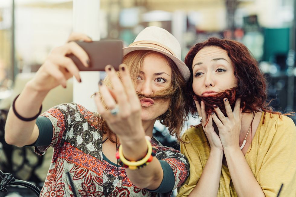 two girlfriends holding hair over faces to look like they have beard and mustache and taking a selfie of it