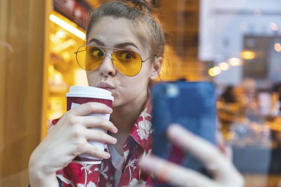 young woman outside in sunglasses taking a selfie while drinking a to go coffee