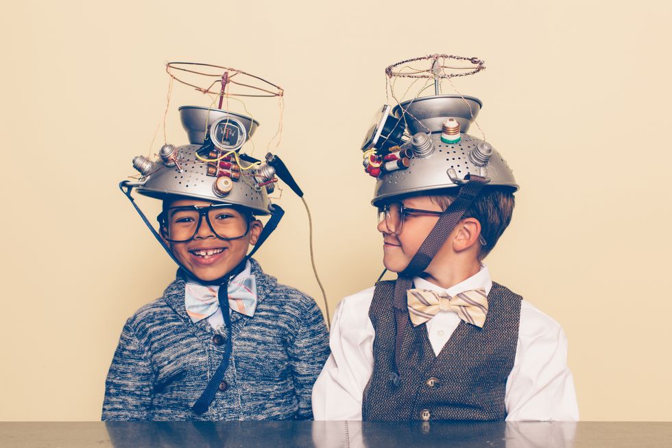 two nerd boys dressed in casual clothing, glasses and bow ties experiment with a homemade science project they are both smiling and sitting at a table, and one is looking at the other with helmets on their heads in front of a beige background retro styling