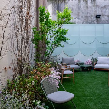 un giardino arredato con sullo sfondo una quinta con pannelli azzurri, un salottino con poltroncine in legno e tavolo in alluminio e una poltrona con schienale in corda