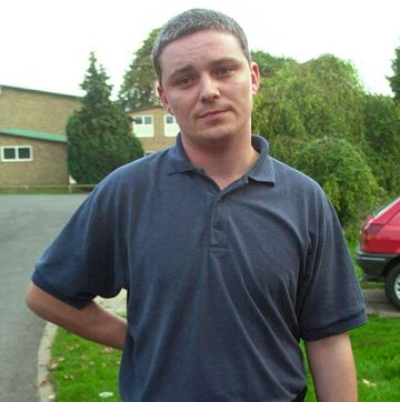 ian huntley stares into the camera whiile standing outside near a lawn, driveway and houses, he wears a blue polo shirt and has one arm behind his back
