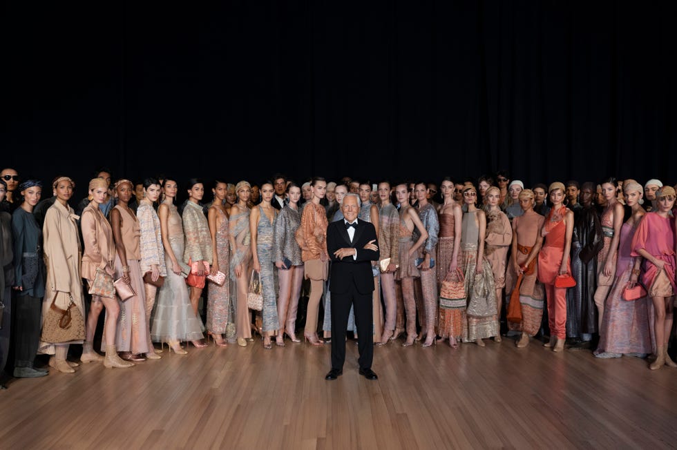 a group of models wearing various fashion outfits stands in formation with an individual in a tuxedo at the center