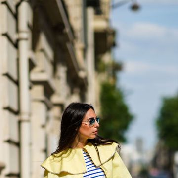a woman wearing a blue and white striped long sleeve shirt with yellow pants, a woven straw tote bag by loewe, and striped slide sandals on a cobblestone street in a roundup of the best beach bags beach totes 2024