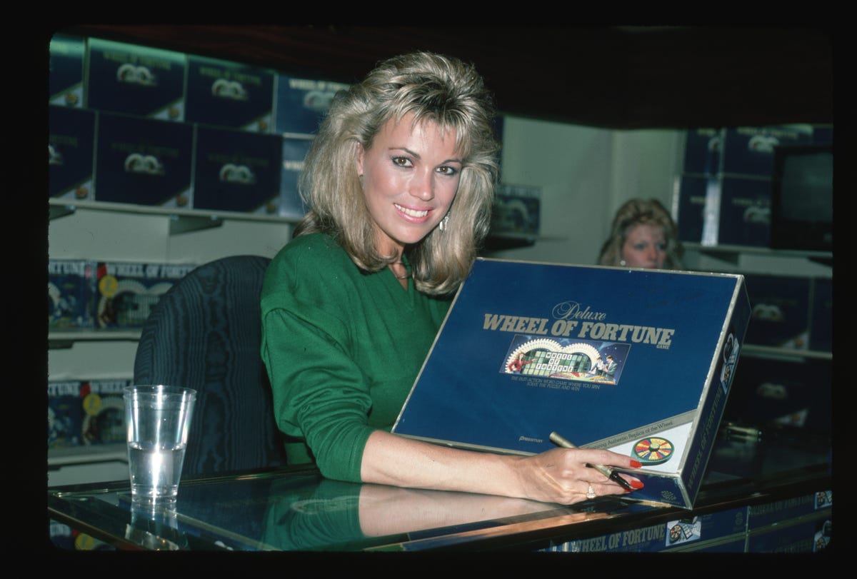 a smiling vanna white poses for the camera while displaying a deluxe wheel of fortune board game, she sits in a desk chair at a table and also holds a marker in her visible right hand that rests below the board game box, she is wearing a kelly green long sleeve shirt with the sleeve rolled up to her elbow, her blonde hair is shoulder length and she has bangs, in the background you can faintly make out other board game boxes as well as the head of another woman sitting behind vanna