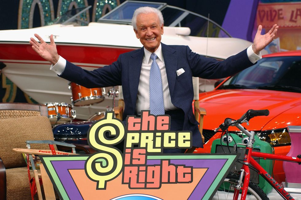 bob barker sits in a directors style chair and opens his arms wide, he is wearing a blue suit and tie and is surrounded by products including a speed boat, red car, drum kit, red bicycle, and snow sled, in front of him is a multicolored sign that says the price is right