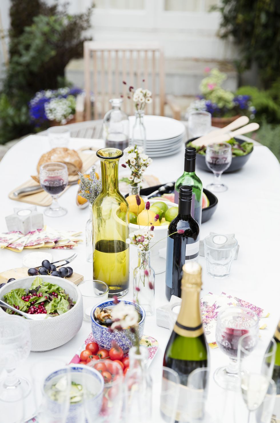 garden party lunch on patio table with wine bottles, a bowl of lemons and limes, grapes, bread and salad