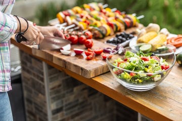 garden table full of food ready for a barbeque party with a salad in the front and a woman cutting vegetable aside