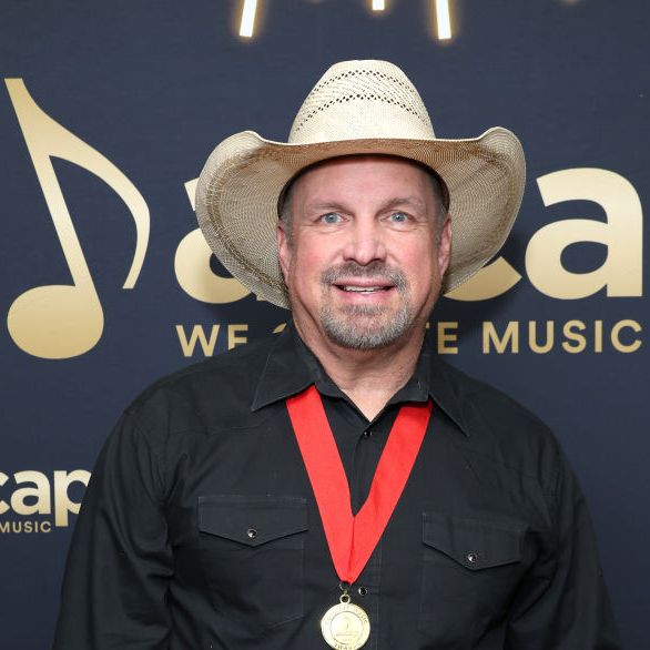 garth brooks smiles at the camera, he wears a straw cowboy hat, black collared shirt and a gold medal with a ribbon lanyard