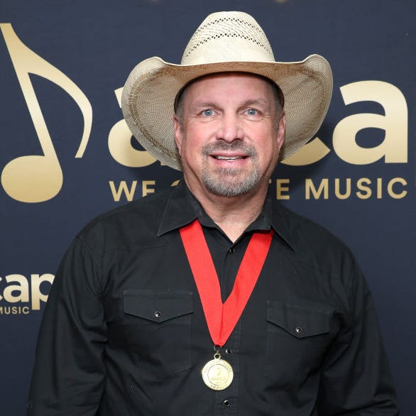 garth brooks smiles at the camera, he wears a straw cowboy hat, black collared shirt and a gold medal with a ribbon lanyard