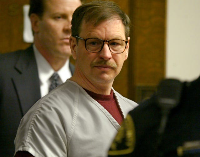 gary ridgway looks to the right of the camera, he wears a gray inmate uniform shirt over a red sweatshirt and tortoise shell glasses, a man in a suit stands behind him