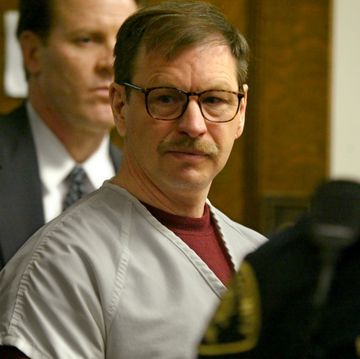 gary ridgway looks to the right of the camera, he wears a gray inmate uniform shirt over a red sweatshirt and tortoise shell glasses, a man in a suit stands behind him