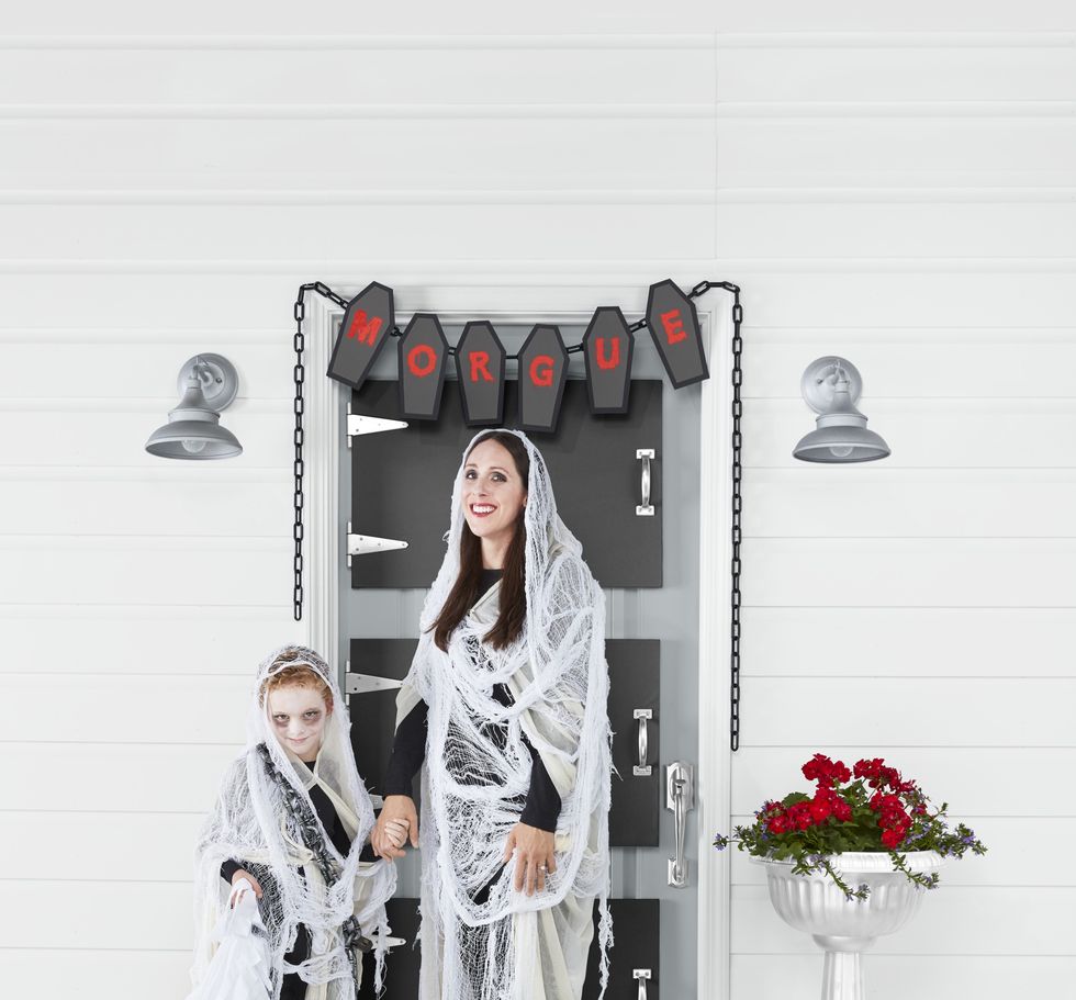 mother and son in ghost family costume standing in front of front door decorated like morgue freezer door for halloween
