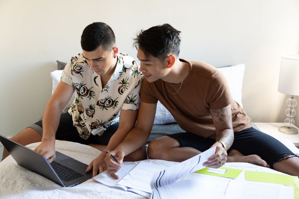 a gay couple sit on their bed with their laptop and paperwork