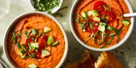 tomato gazpacho in a bowl topped with chopped cucumbers, basil, sliced cherry tomatoes, and an olive oil drizzle