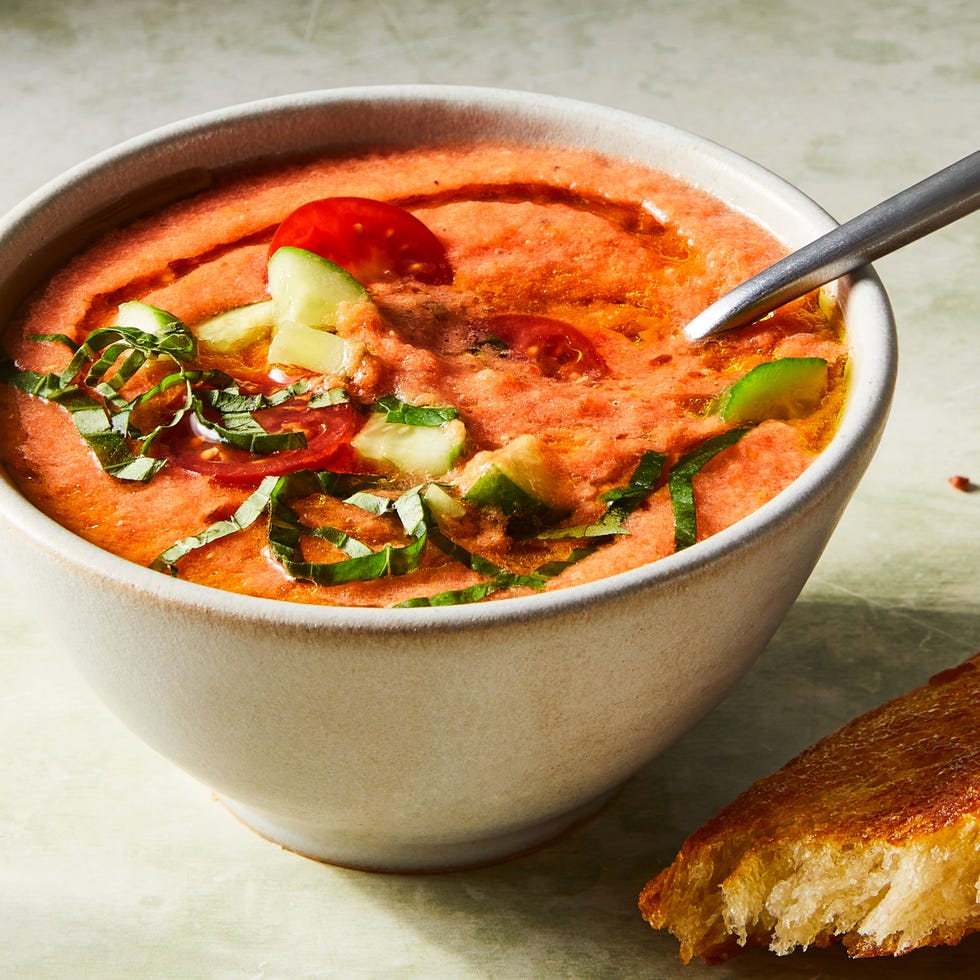 tomato gazpacho in a bowl topped with chopped cucumbers, basil, sliced cherry tomatoes, and an olive oil drizzle