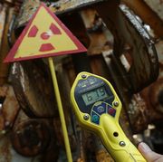 yellow digital geiger counter in the foreground with rusty metal and a nuclear warning sign in the background