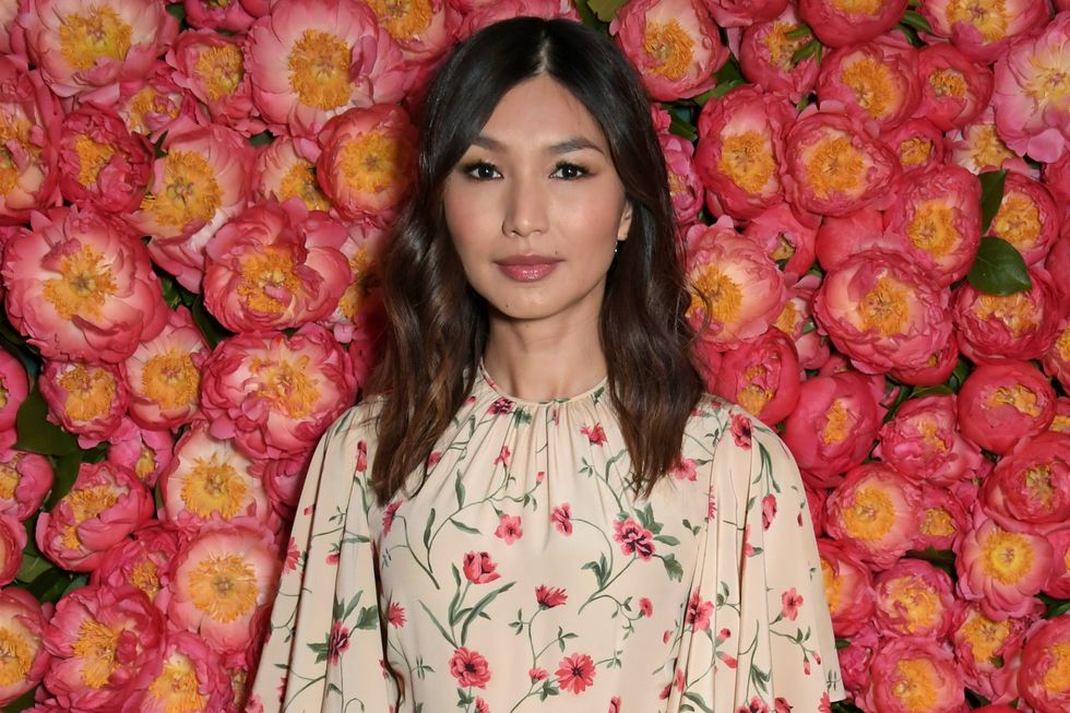 gemma chan wearing a cream floral dress and standing in front of a pink flower wall