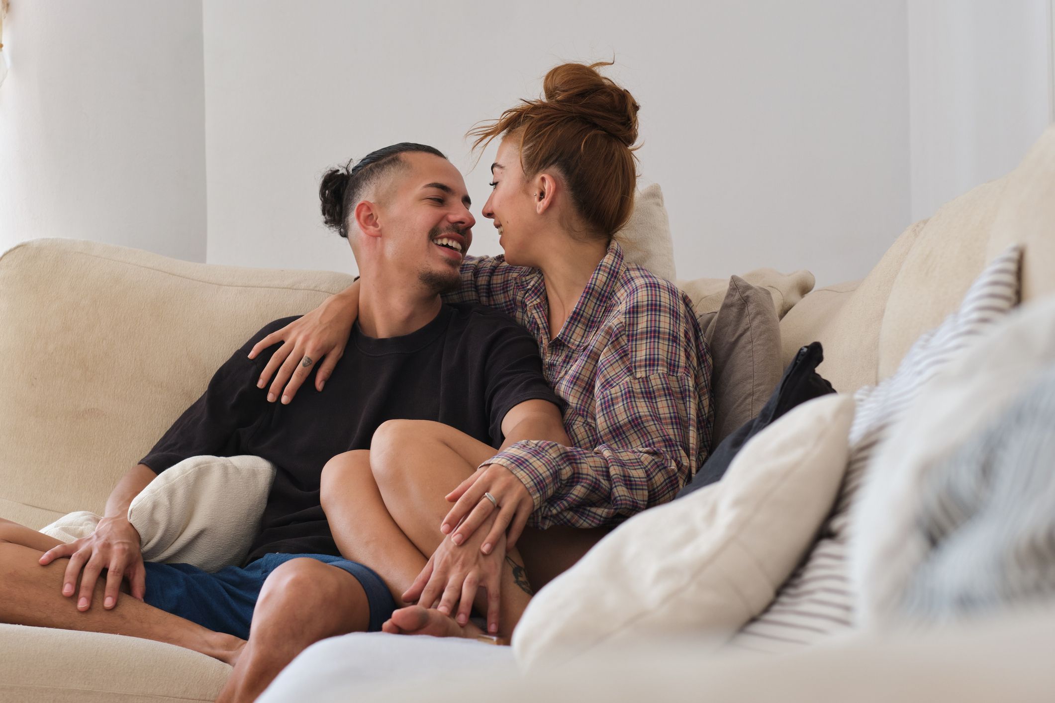 general shot of a latino young female and male couple looking at each other, joking and smiling sitting on the sofa