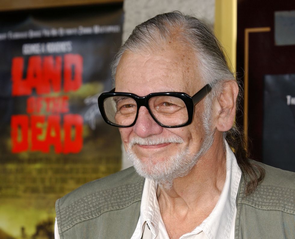 george romero looks off camera and smiles, wearing a white shirt, tan vest, and large black rimmed glasses
