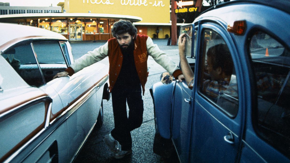 George Lucas posing between two cars