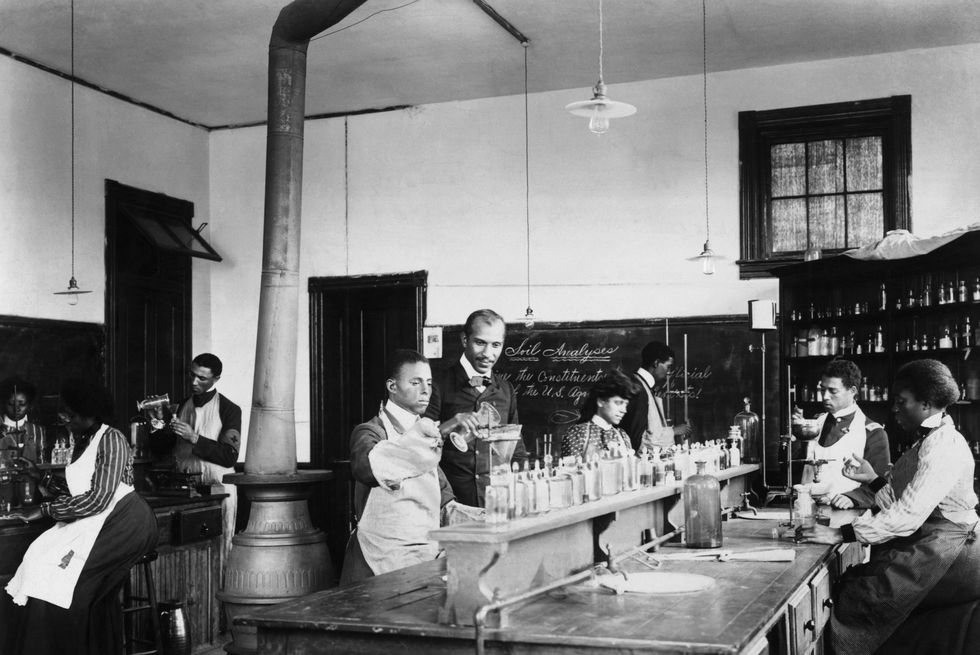 george washington carver watches a student pour a substance into another vessel as several other students sit at lab tables nearby and work