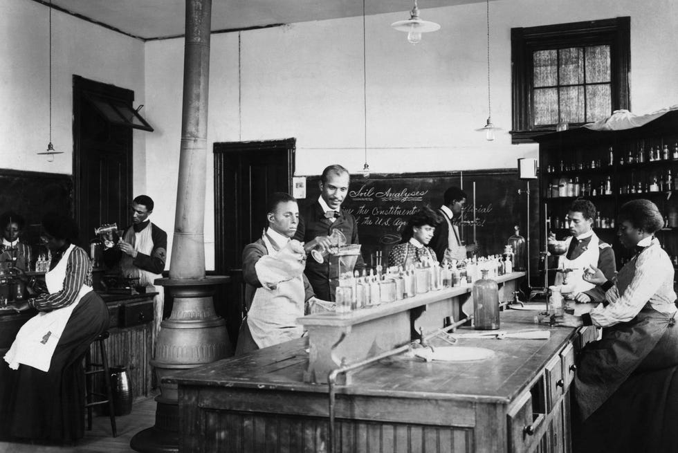 George Washington Carver observa a un estudiante verter una sustancia en otro recipiente mientras varios otros estudiantes se sientan en mesas de laboratorio cercanas y trabajan.