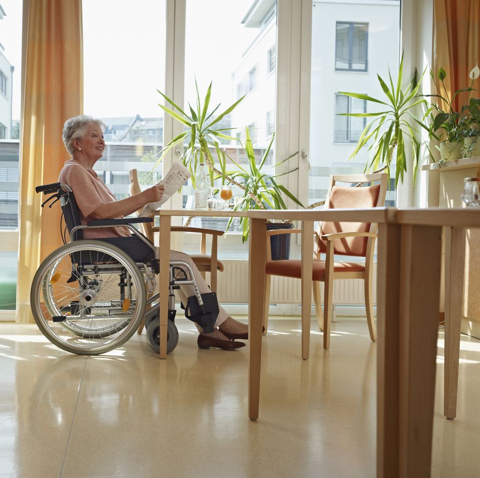 a senior woman in a wheelchair reading a newspaper in a sunny room with a wooden table and plants