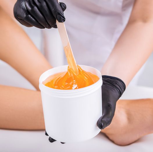 a beauty therapist holding a pot of wax with a womans legs in the background