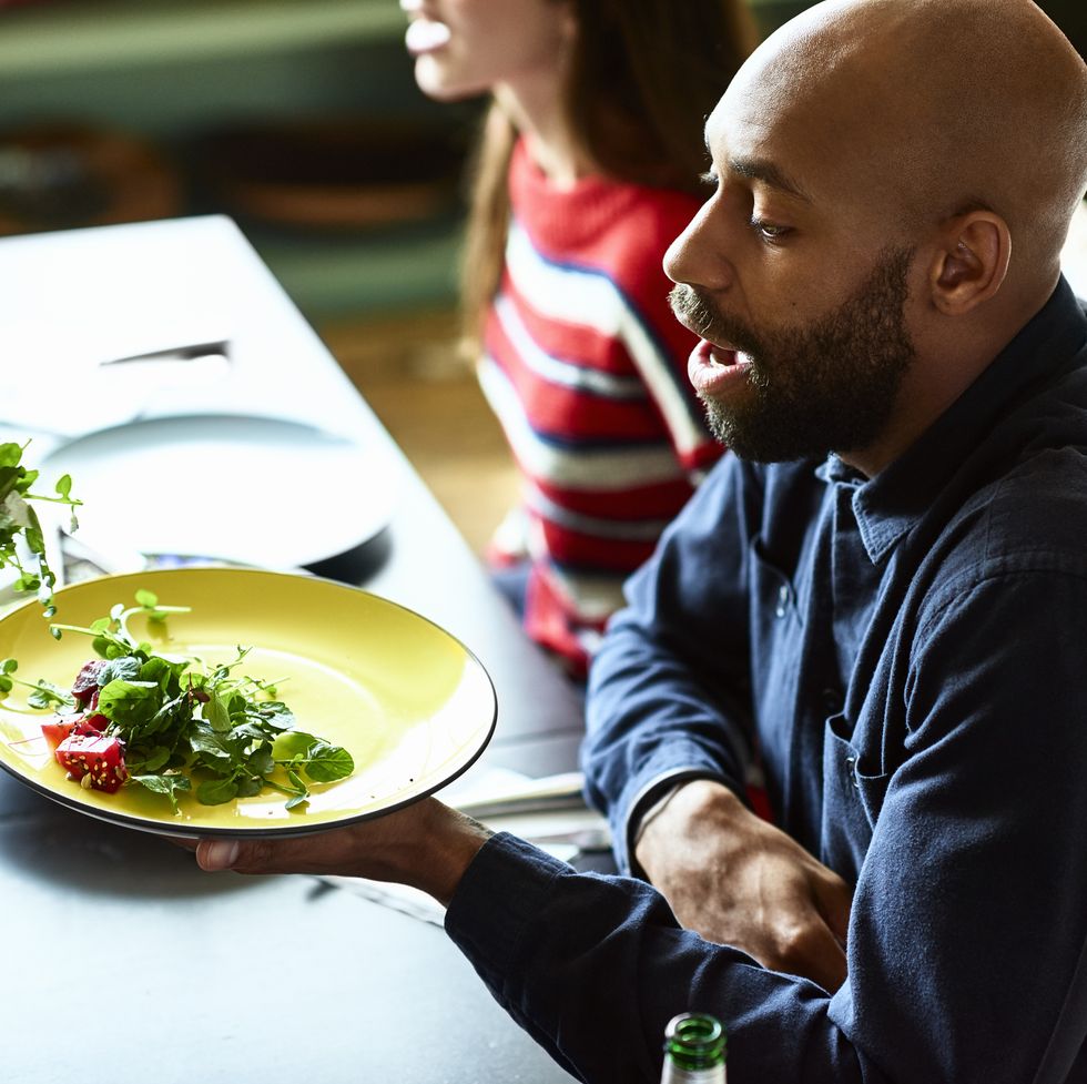attractive hipster african american man sitting at dining table with friends, talking with serious expression, wearing casual blue shirt with a shaved head and beard