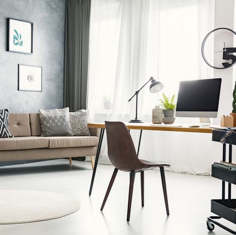 Real photo of a bright home office interior with a sofa, graphics, desk with a computer and lamp and modern clock