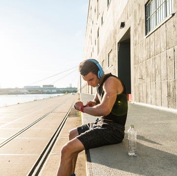 Young athlete wearing headphones, sitting on a wall, checking smartwatch Individual in sportswear training while seated by a waterfront