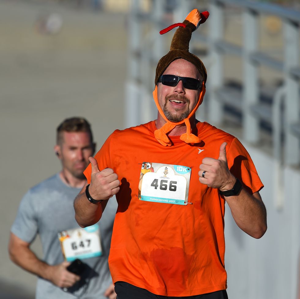 a white man running in a turkey trot wearing an orange tshirt and a turkey hat giving a double thumbs up
