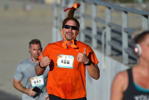 a white man running in a turkey trot wearing an orange tshirt and a turkey hat giving a double thumbs up