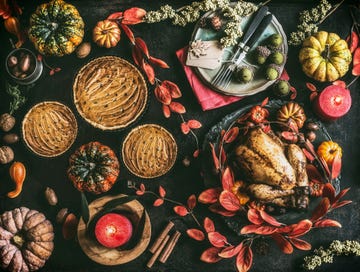 thanksgiving day dinner table with roasted whole chicken or little turkey , pumpkin pie on dark rustic background with plates, sauce, cutlery, table setting tags, burning candles, red autumn leaves, nuts and festive decoration , top view, still life, flat lay