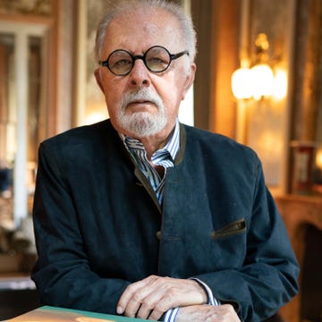 fernando botero stares at the camera with a neutral expression on his face, he wears round black glasses and a navy suede jacket over a blue and white striped collared shirt, his hands are crossed in front of him as he leans slightly left