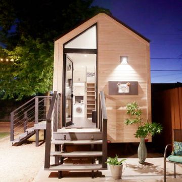 a tiny house with large glass windows, sits in the backyard at night, surrounded by trees and party lights