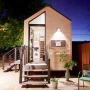 a tiny house with large glass windows, sits in the backyard at night, surrounded by trees and party lights