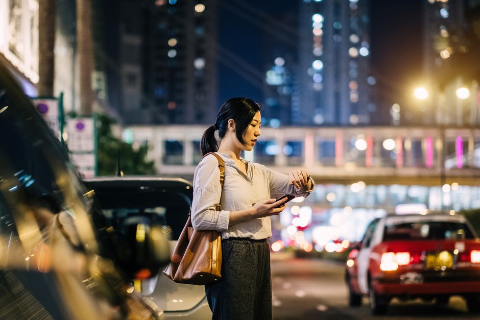 young woman with mobile phone checking time on wristwatch by busy street in city at night