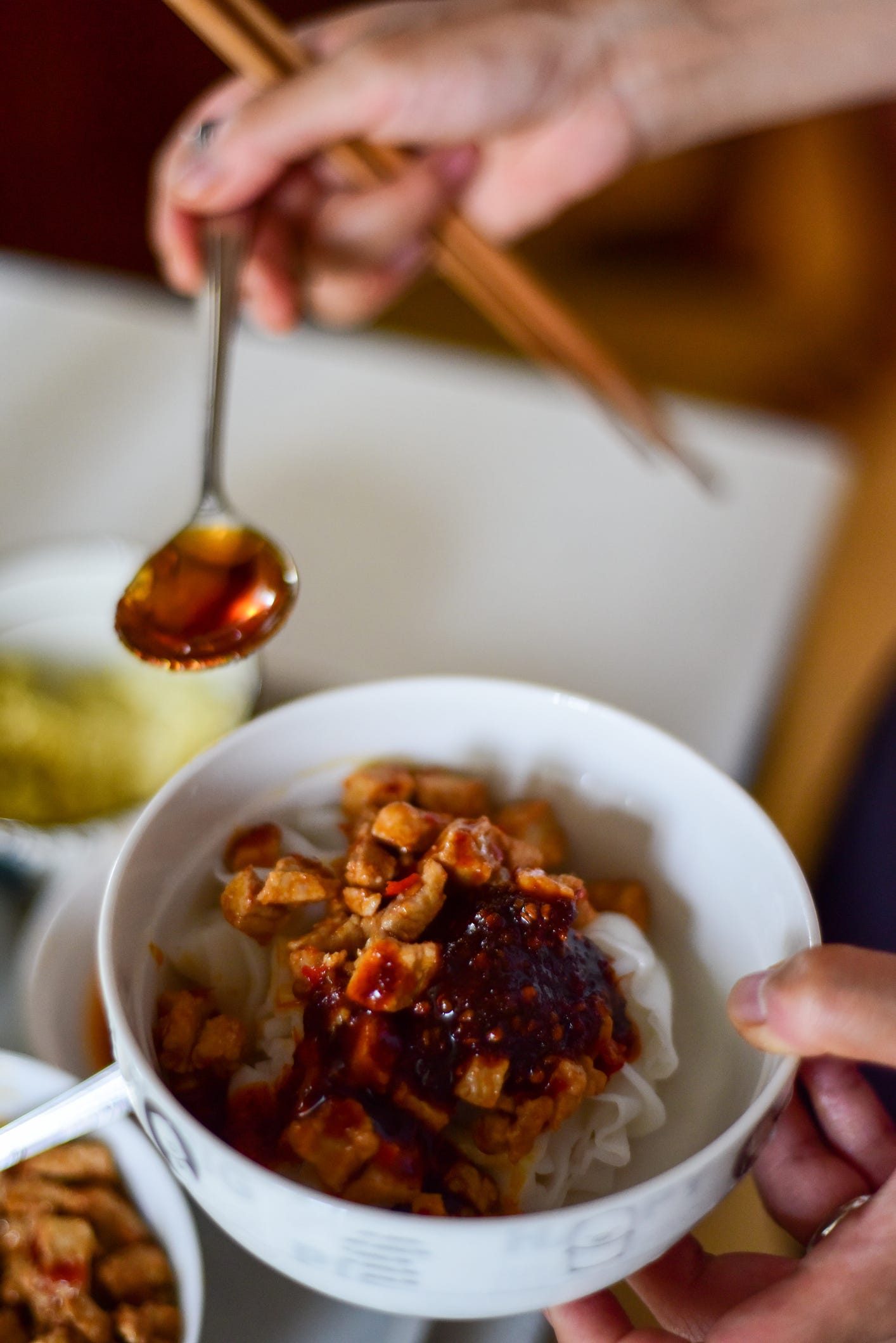 Adding chili oil to spicy pork and freshly made rice noodles in a bowl using a spoon