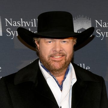 toby keith smiles at the camera, he wears a black cowboy hat, black coat, and white collared shirt with a navy bandana tied around his neck, he stands in front of a gray background with white writing