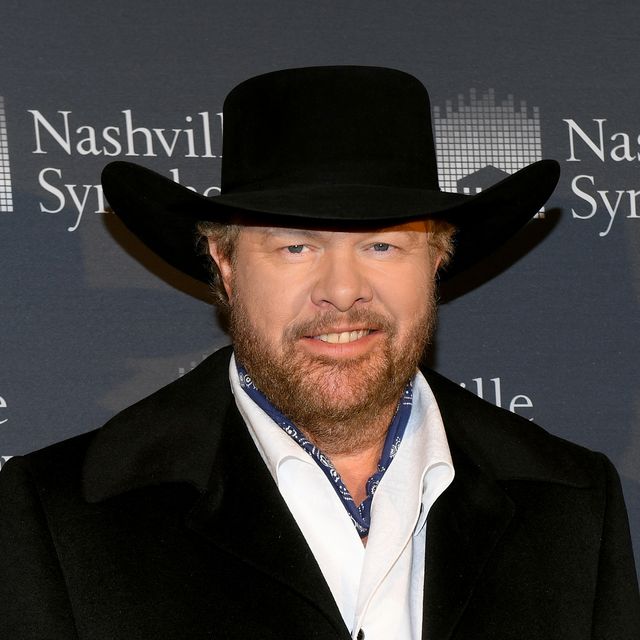 toby keith smiles at the camera, he wears a black cowboy hat, black coat, and white collared shirt with a navy bandana tied around his neck, he stands in front of a gray background with white writing