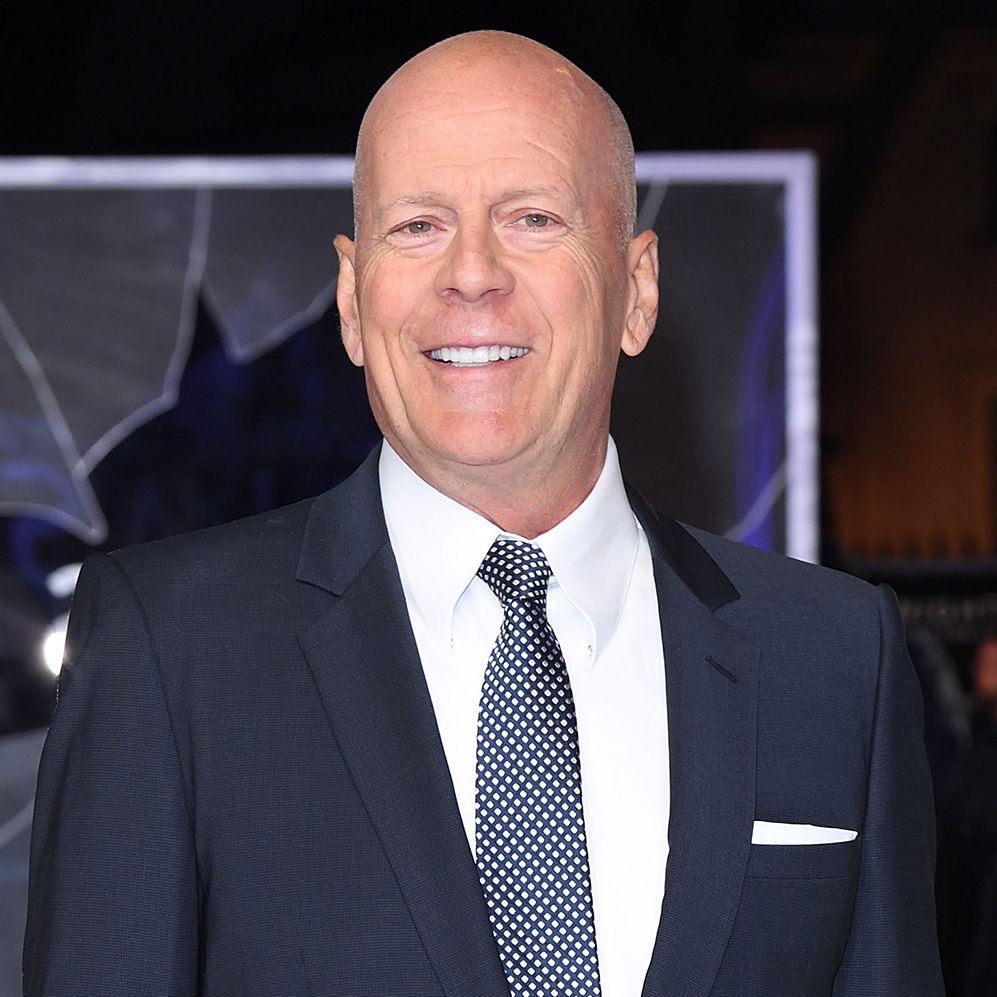 bruce willis smiles at the camera while wearing a navy suit jacket, white collard shirt, and navy blue tie with white polka dots, he has a white pocket square in his jacket breast pocket and he is bald