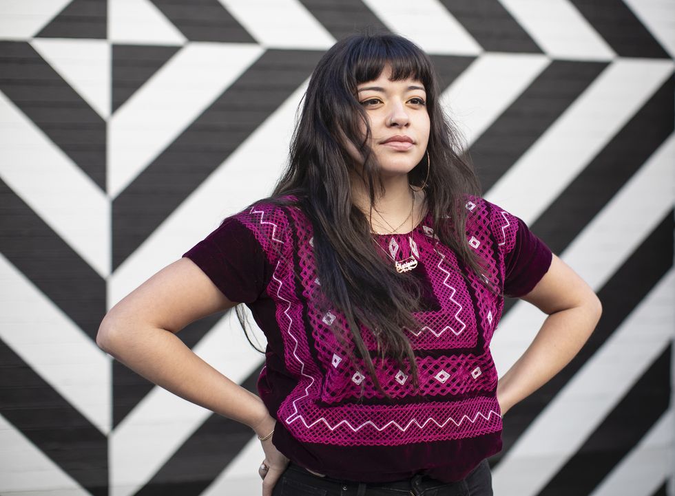 millennial latina stands in front of a wall painted with vivid black and white stripes she wears a magenta woven textile blouse that echoes the geometry of the wall behind her long dark hair and deep brown eyes, muscular build and clear complexion reflect her natural beauty and vitality with hands on hips, we see gold bracelet and gold necklace and hoop earring she has a quiet and confident smile, looking just to the side of the camera, waiting for a friend or looking at something that inspires her