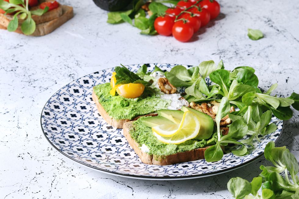 Vegetarian sandwiches with avocado, ricotta, egg yolk, spinach, cherry tomatoes on whole grain toast bread on ceramic plate with ingredients above over white marble kitchen table Close up