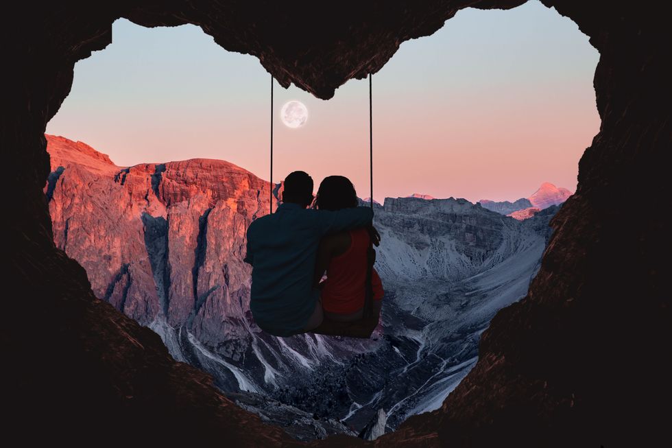 composition of the alps mountains during sunset with full moon and couple on swing from a heart shape cave