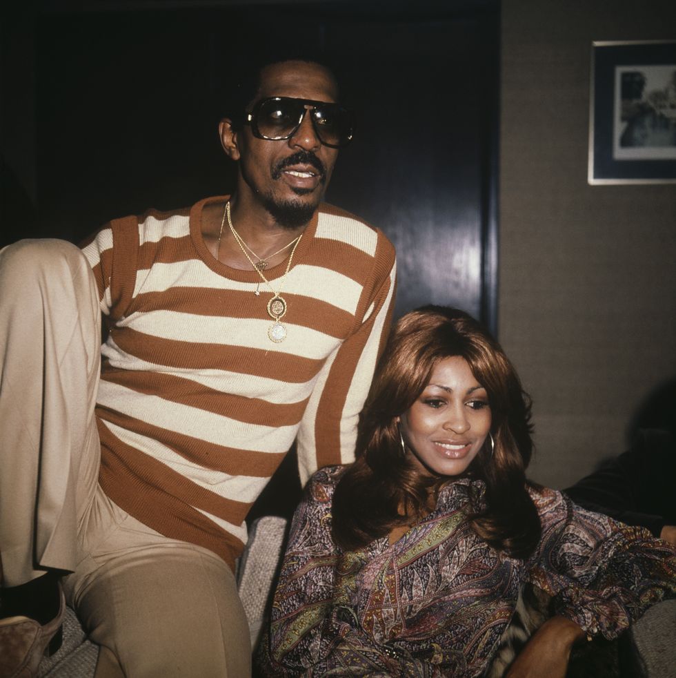 ike turner and tina turner sit on a couch next to each other, ike wears an orange and white striped shirt with khakis, gold necklaces and sunglasses, tina wears a paisley top and hoop earrings