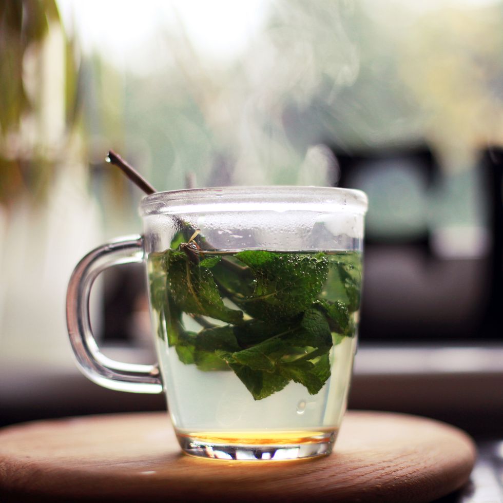 fresh mint tea, with mint leaves and honey, in a glass cup stood on wooden board by a kitchen window plant in the background a cafe sign is also in background, partly covered