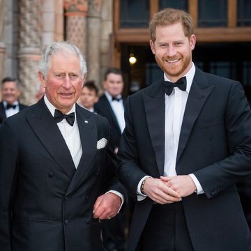 king charles and prince harry stand next to each other and smile, both men wear suit jackets, bow ties, and white collared shirts